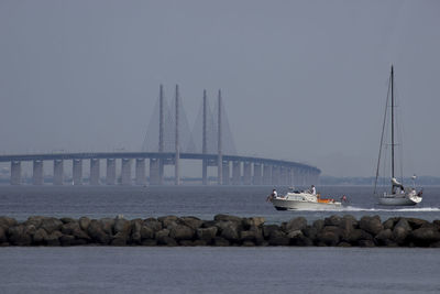 Boats sailing by bridge in sea against sky