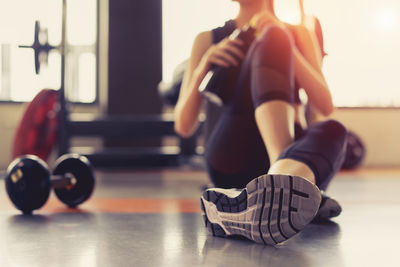 Low section of woman holding water bottle while sitting on floor in gym