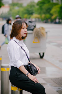 Side view of young woman sitting on bollard by street at city