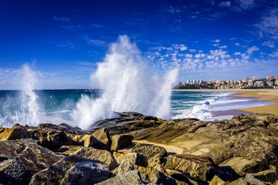 Waves splashing on rocks at shore against blue sky