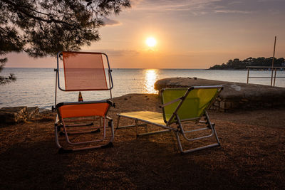 Empty chairs and tables by sea against sky during sunset