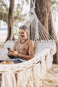 Smiling shirtless man using smart phone while sitting on hammock at beach
