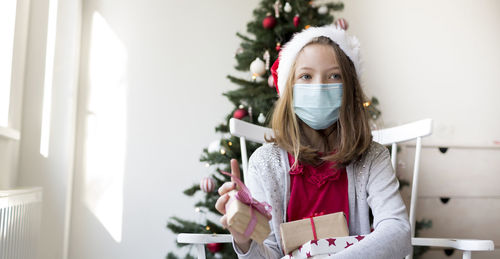 Girl wearing mask sitting against christmas tree at home