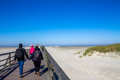 Rear view of people walking on pier at beach against clear blue sky during sunny day