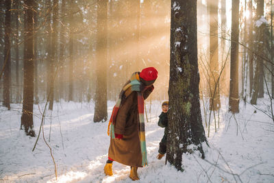 Mother in coat, colorful scarf and red hat playing with her son hide and seek in sunny winter day. 