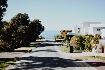 Empty road by houses leading towards sea against clear sky