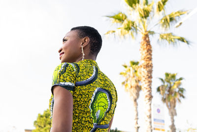 From below of confident happy young african female with short dark hair in bright summer dress looking away while standing near palms on city street on sunny day