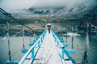 Rear view of man walking snowcapped footbridge over river during winter