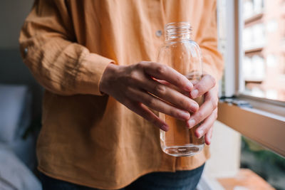 Midsection of young woman holding water bottle by glass window at home