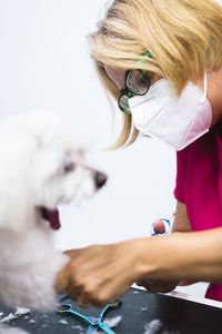 Crop anonymous female groomer in respiratory mask and eyeglasses cutting fur of maltese dog on grooming table in salon during covid 19 pandemic