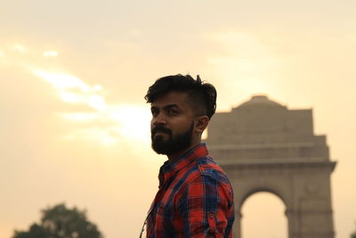 Thoughtful man with triumphal arch against cloudy sky during sunset