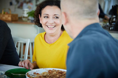 Cheerful mature woman talking with male friend while having lunch at home