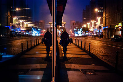 Rear view of woman walking on sidewalk reflecting on window at night