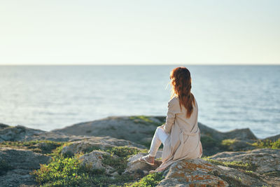 Woman sitting on rock looking at sea against clear sky