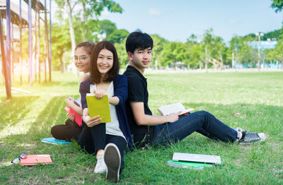 Portrait of friends studying while sitting on grassy field against sky at park