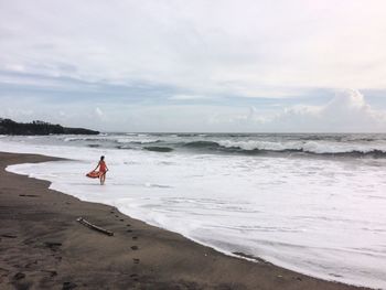 Woman holding scarf while standing at shore against cloudy sky