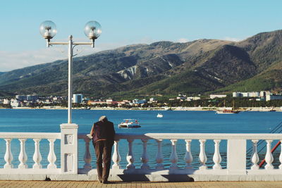 Man standing on railing by sea against mountains