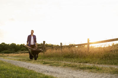 Man pushing wheelbarrow on dirt road by fence against clear sky