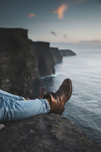 Low section of person wearing shoes on cliff by sea during sunset