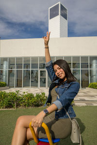 Woman gesturing peace sign sitting against building