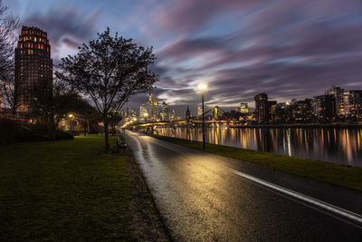 Illuminated street by river and buildings against sky at dusk