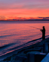 Man fishing in sea against sky during sunset