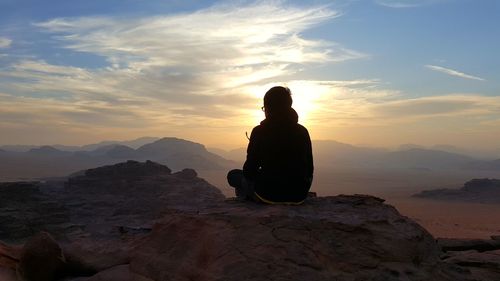 Boy sitting on rock formation against sky during sunset