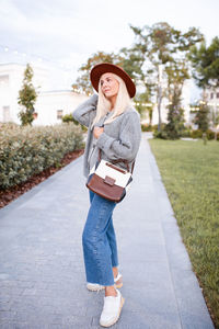 Smiling beautiful blonde teenage girl 17-18 year old wear stylish hat, bag, knit gray jumper outdoor