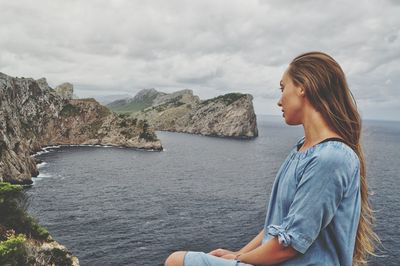 Side view of woman sitting on rock by sea against sky