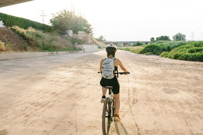 Back view confident young female bicyclist in sportswear and helmet riding bike on fenced curvy paved track while training alone in summer day