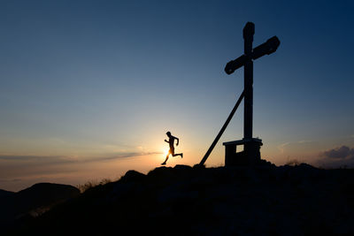 Silhouette of man running in the setting sun on top of a mountain with the cross