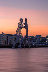 Molecule men sculptures in spree river against sky during sunset