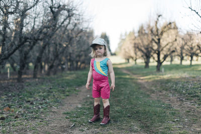 A young girl stands in an orchard wearing a leotard and cowgirl boots.