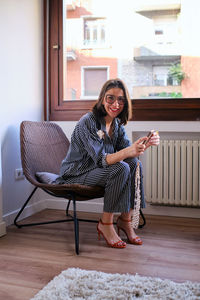 Full body side view of female in eyeglasses and high heels text messaging on cellphone while sitting on armchair near window