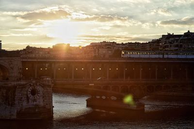 Train on railway bridge over river in city against sky during sunset