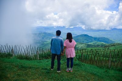 Rear view of man and woman holding hands while standing on grass against sky