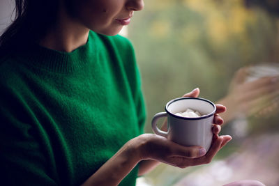 Woman in a green sweater holds a metal mug of cocoa and marshmallows