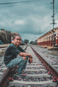 Portrait of man sitting on railroad track against cloudy sky