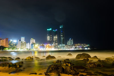 Illuminated buildings by sea against sky at night