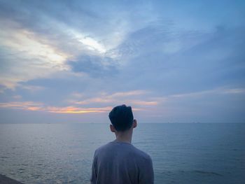 Rear view of young man looking at sea while standing against cloudy sky during sunset