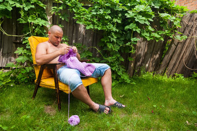 A young bald man sits on an armchair and knits a sweater from natural lilac wool threads 