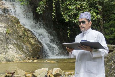 Man reading book while standing against waterfall