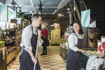 Smiling sales clerk holding broom while looking at saleswoman in supermarket