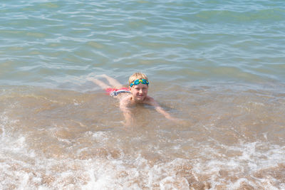 Boy 8 years old in swimming goggles lies in the surf on the sandy beach of the blue sea, loves to
