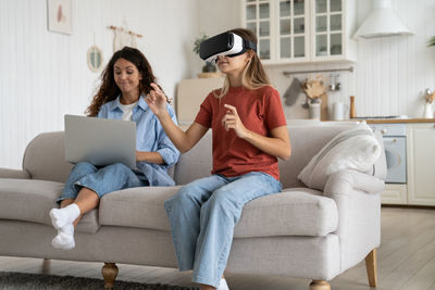 Concentrated little teen girl with vr glasses visits 3d metaverse and sits on sofa near mother