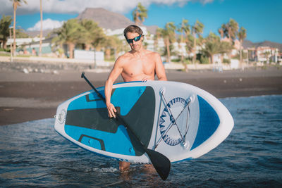 A young sportsman man practices paddle surfing on the beach under a blue sky