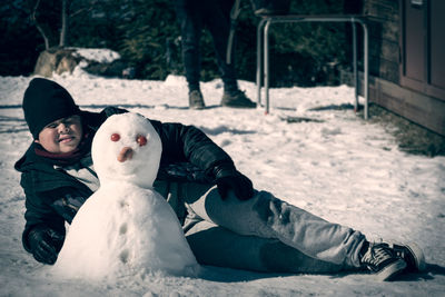 Boy lying in the snow next to self-made snowman with carrot nose and tomato eyes,