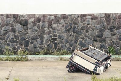 Abandoned sofa on footpath against stone wall