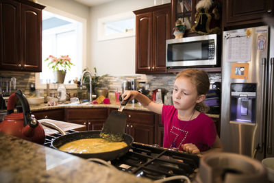 Candid image of unsmiling young girl cooking eggs in messy kitchen