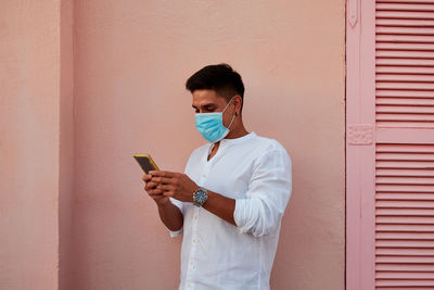 Young latin man with mask is looking his phone on pink background
